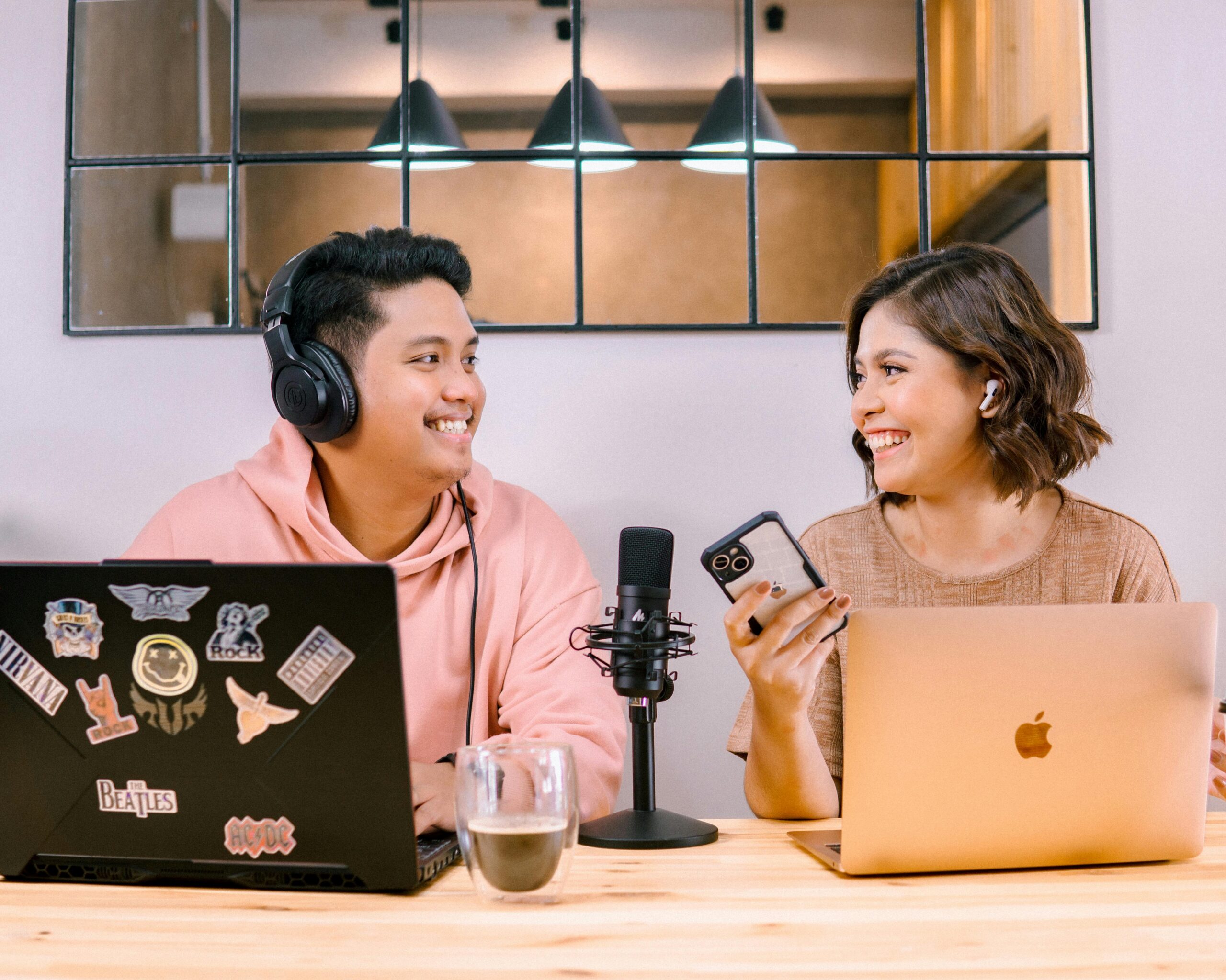 Two adults recording podcast at home, smiling and interacting with laptops and microphone.